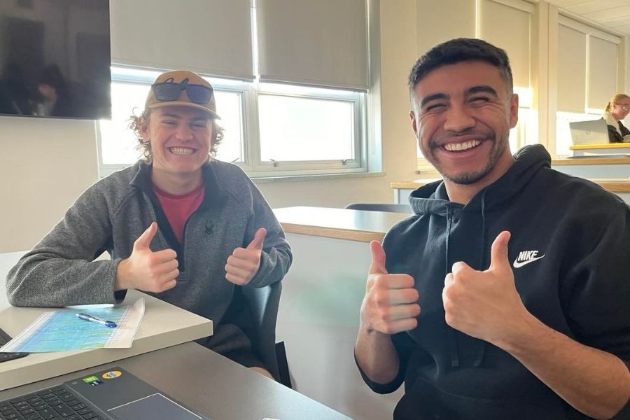 Two students sit at a desk in the classroom smiling and giving thumbs up for the photo