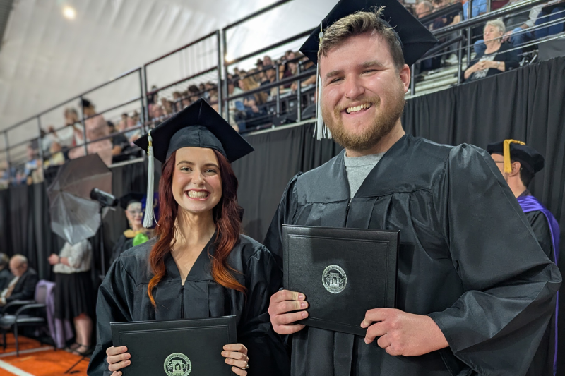 two students in cap and gown hold diplomas
