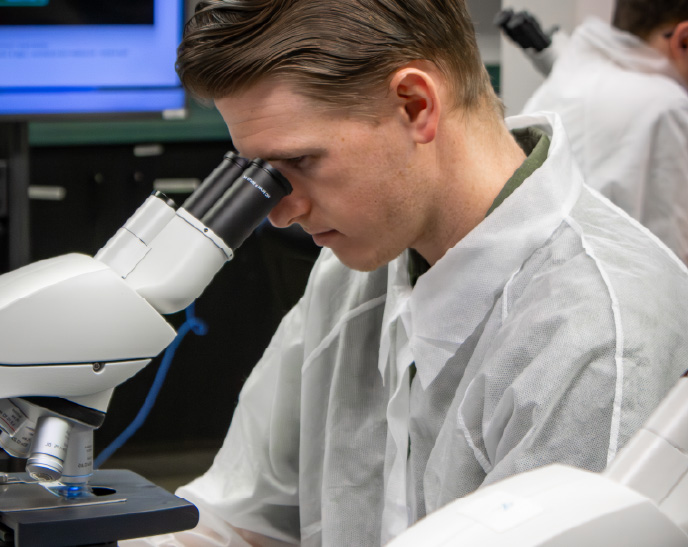 A student wearing a lab coat looks through a microscope during a science laboratory class. Additional microscopes and students are visible in the background, highlighting a hands-on learning environment.