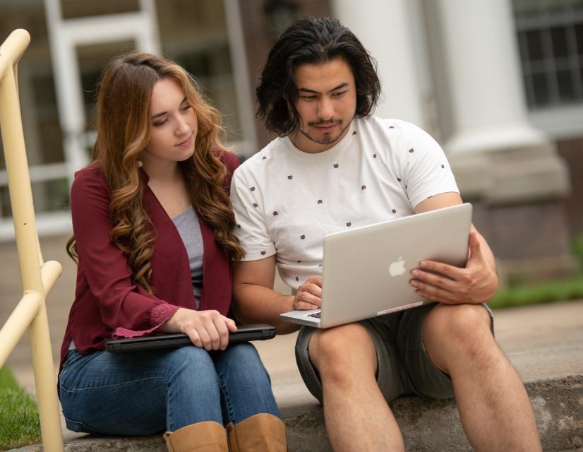 Two students sitting on steps in front of building viewing laptop.
