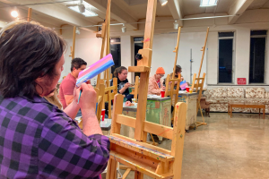 Students stand at easels in a studio room and paint on small canvases