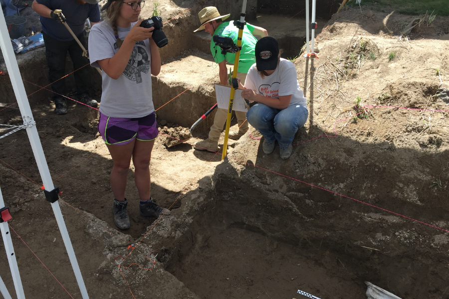 Students are digging in the dirt outside. One student holds a camera and is taking a photo. Another student is making notes on a paper.