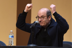 David Grann sits at a table with a microphone in front of him. His arms are upraised in a gesture