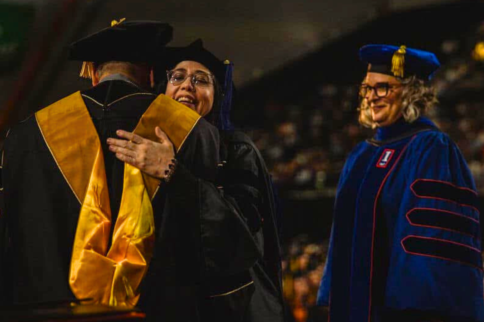 Three individuals in academic regalia, two hugging and one smiling beside them.