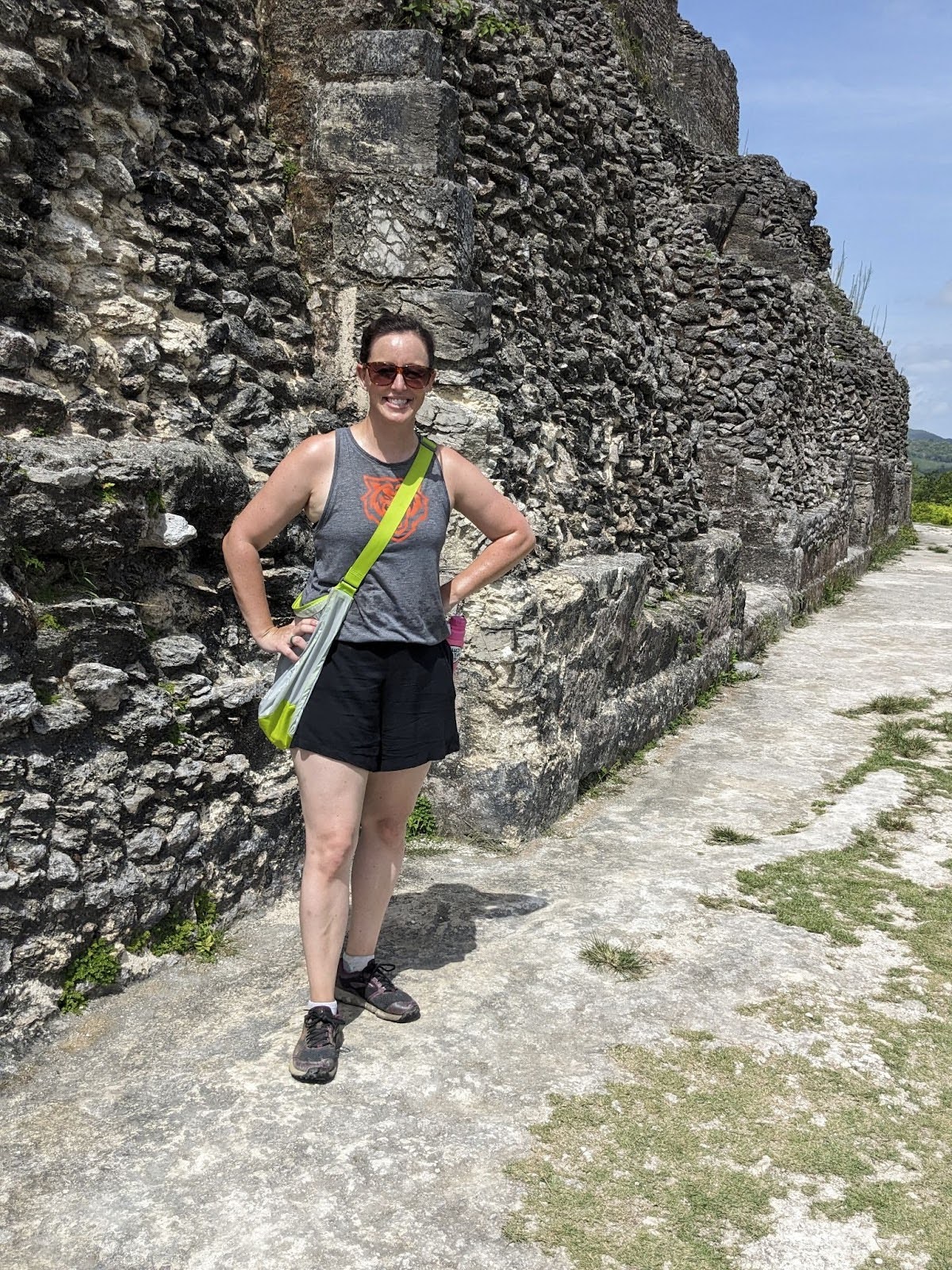 A woman in casual attire stands next to an ancient stone wall under a clear blue sky.