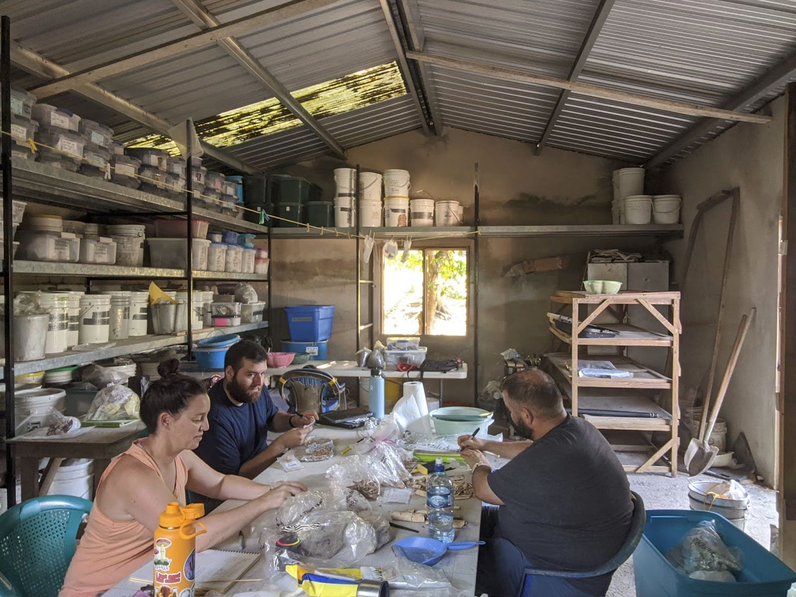 Three people working at a table inside a shed filled with storage shelves and containers.