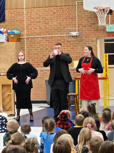 Three performers entertain children in a school gym with a brick wall backdrop.