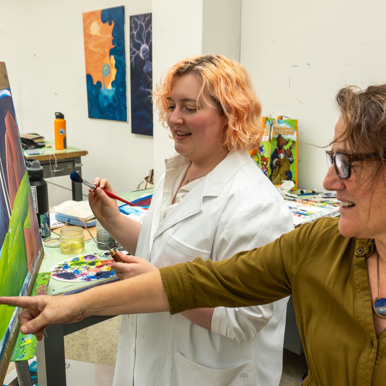 A student holds a paintbrush at a canvas on an easel while a professor points to something on the canvas