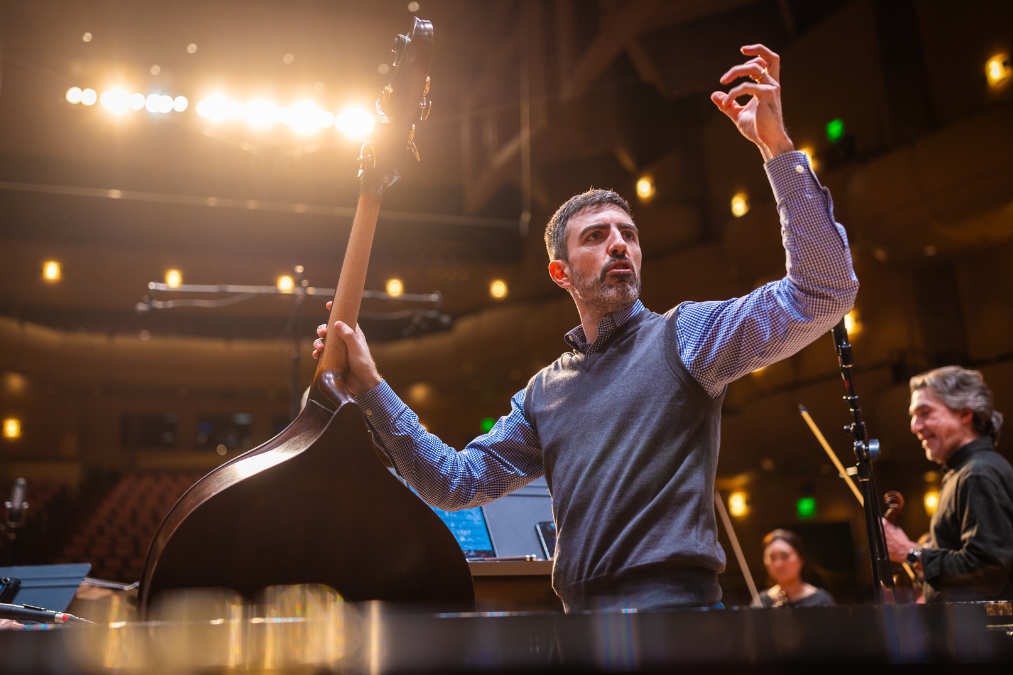 Pedro Giraudo stands onstage backlit by stage lights. He is raising his arms in conducting motion