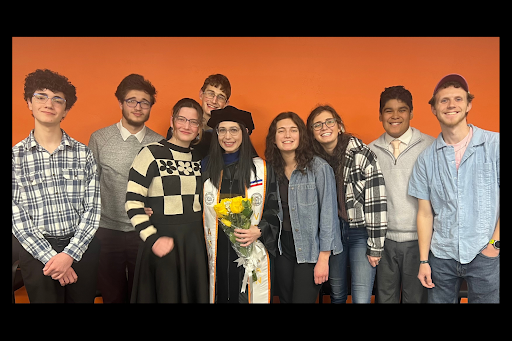 A group of nine people poses in front of an orange background. Mel wears graduation attire and holds yellow flowers.