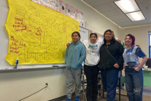 Luzahan Matsaw, Aiyana Eschief and Seth Stacey stand next to their debate coach, Abbey Vaughn, and a poster wishing them luck at the National Speech & Debate Tournament.