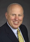 A photo of a bald man with light skin wearing a suit and a yellow tie, smiling.