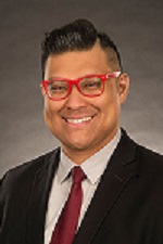A man with medium-dark skin tone and black hair. Wearing red glasses, a black suit jacket, a white button down and a red tie. He is smiling.