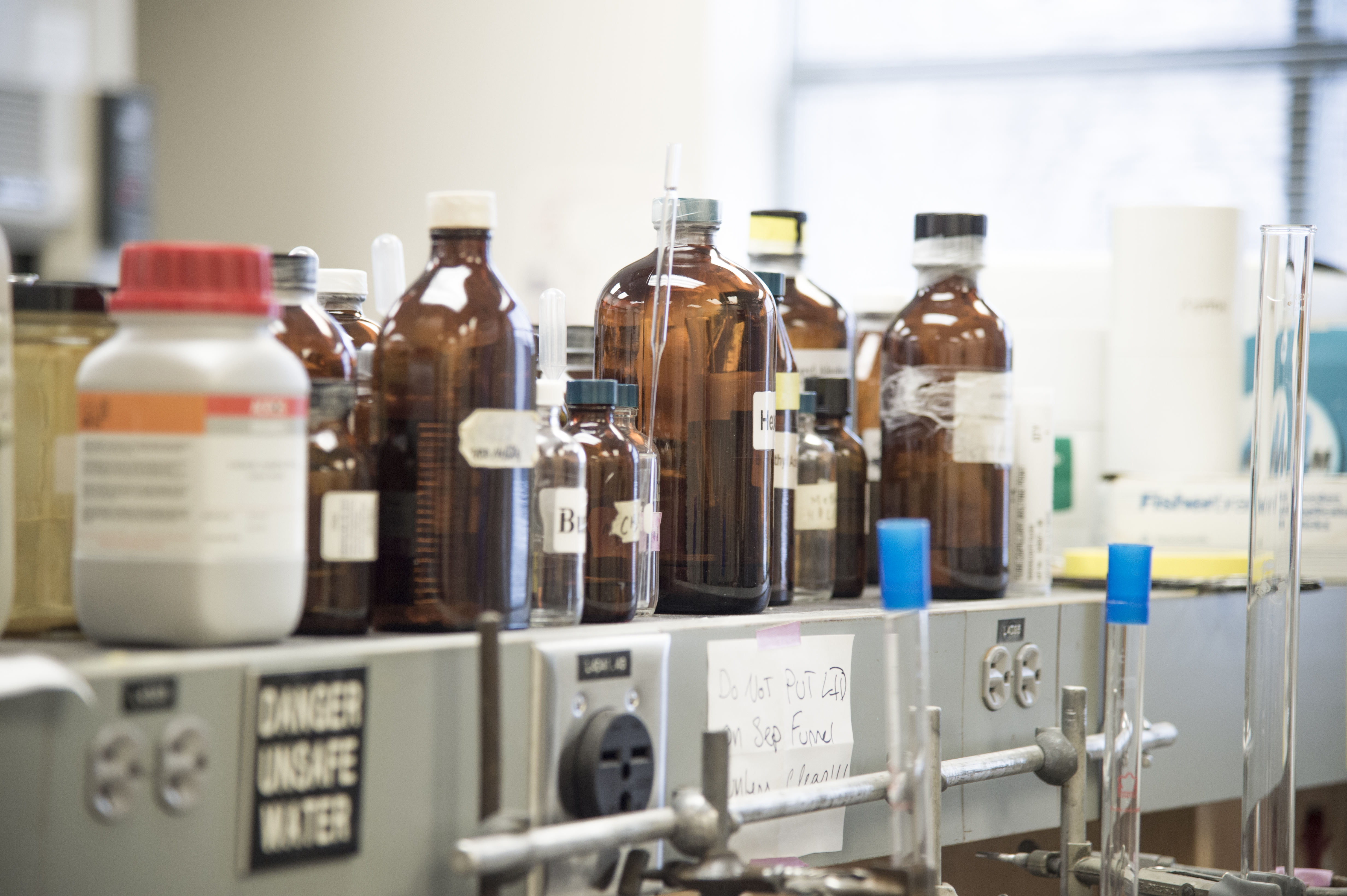 Photo of multiple chemical bottles lined up along the top shelf of a lab workspace.
