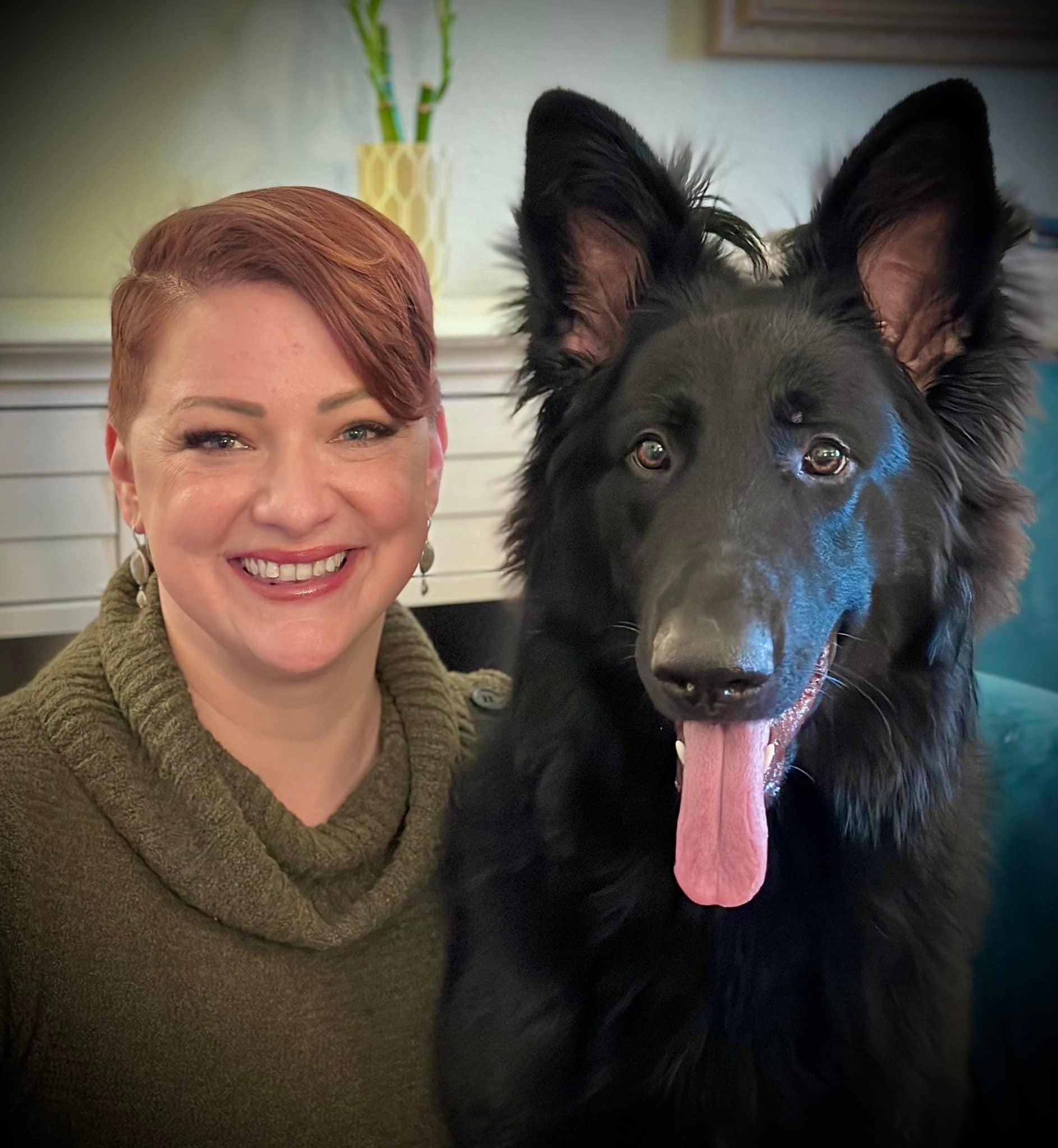 A woman with red hair sitting to the right of a large black german shepard dog. The dogs ears and pointed up, and their tongue is out. Both woman and dog look into the camera.