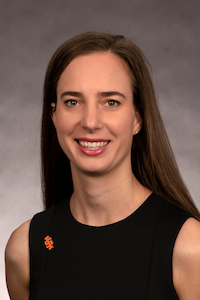 A feminine person with medium skin tone and brown hair. She is wearing a black shirt and smiling towards the camera.