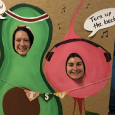 Two women put their heads into cardboard image of avocado (time to avo-cardio) and beet (turn up the beat)