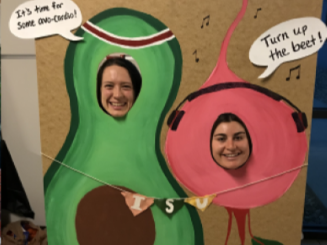 Two women put their heads into cardboard image of avocado (time to avo-cardio) and beet (turn up the beat)