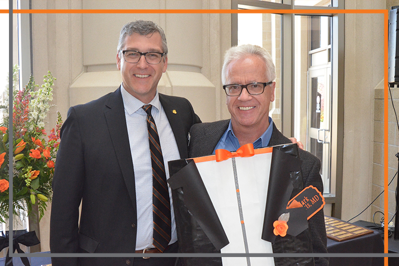 Dr. Mark Roberts holds a tuxedo shaped plaque with Dr. Rex Force in the Stephens Performing Arts Center rotunda