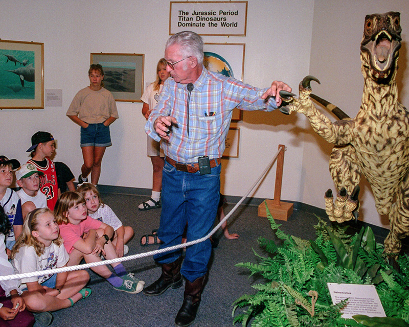 A museum worker displays a model of a dinosaur to a group of children.