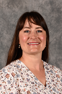 A woman with light skin tone and brown hair. She is wearing a patterned floral shirt and smiling.