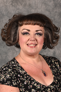 A fair skin toned woman with reddish-purple hair. She is wearing a floral shirt and a necklace. She is smiling.