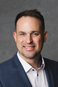 A fair skin tone man with dark, short hair is wearing a white shirt and navy jacket, smiling.