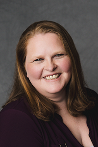 A fair skin tone woman with long auburn hair. She is wearing a black shirt and smiling.