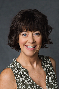 A fair skin toned woman with brown hair and a patterned sleeveless shirt. She is smiling.