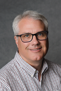 A man with fair skin tone and white hair. He is wearing a black and white checkered button down and black-rimmed glasses.