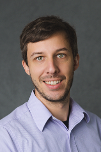 A fair skin tone man with trimmed stubble. He is wearing a lavender shirt and smiling.