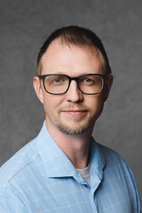 A fair skin tone man with short brown hair and neatly trimmed beard. He wears glasses and a blue collared shirt.