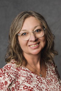 A medium skin toned woman with silver hair. She is wearing glasses and a floral shirt. She is smiling