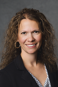 A medium skin tone woman with long, curly dark brown hair. She is wearing a patterned shirt with a black jacket and hoop earrings, smiling.