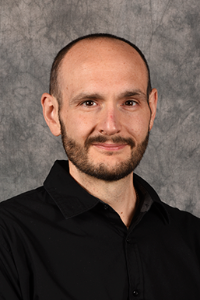 A medium skin toned man with brown hair and a neatly trimmed beard. He is wearing a black button down shirt.