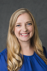 A fair skin tone woman with long blonde hair. She is wearing a blue shirt and is smiling..