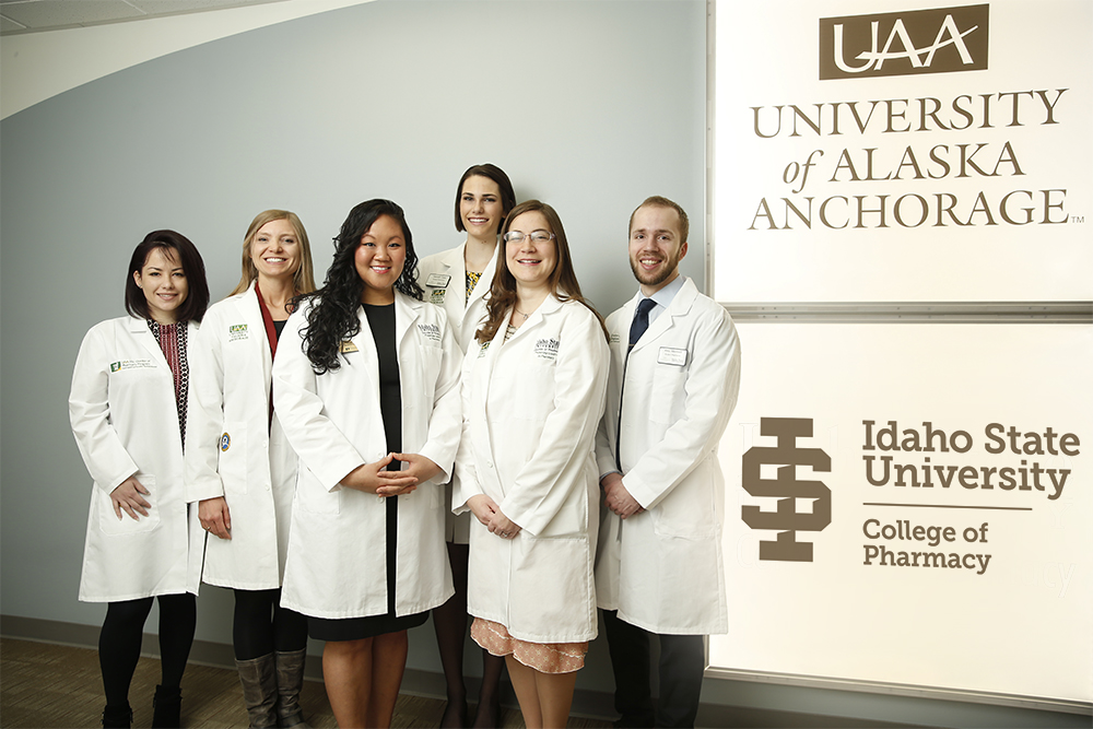 The first six UAA / ISU graduates pose in white coats in front of the UAA/ISU signage at UAA
