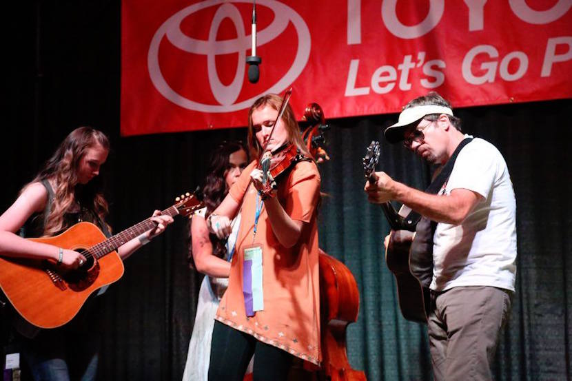 A photo of Murdock playing the fiddle flanked by two guitarists playing music on stage.