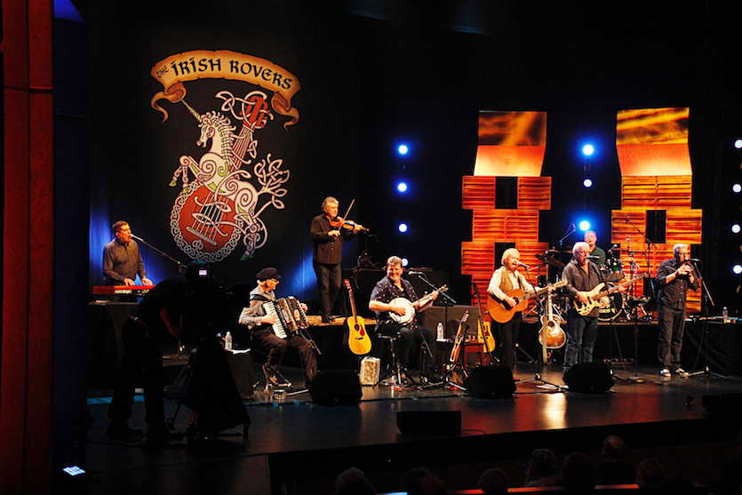 Photo of Irish Rovers on stage in concert, with banner in background.