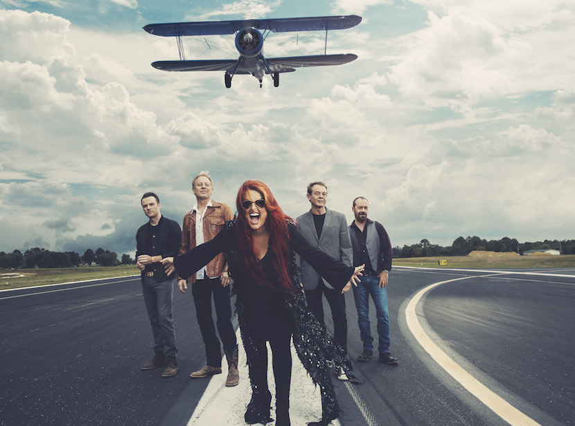Promo photo of Judd with her band standing on a runway with a plane flying overhead.