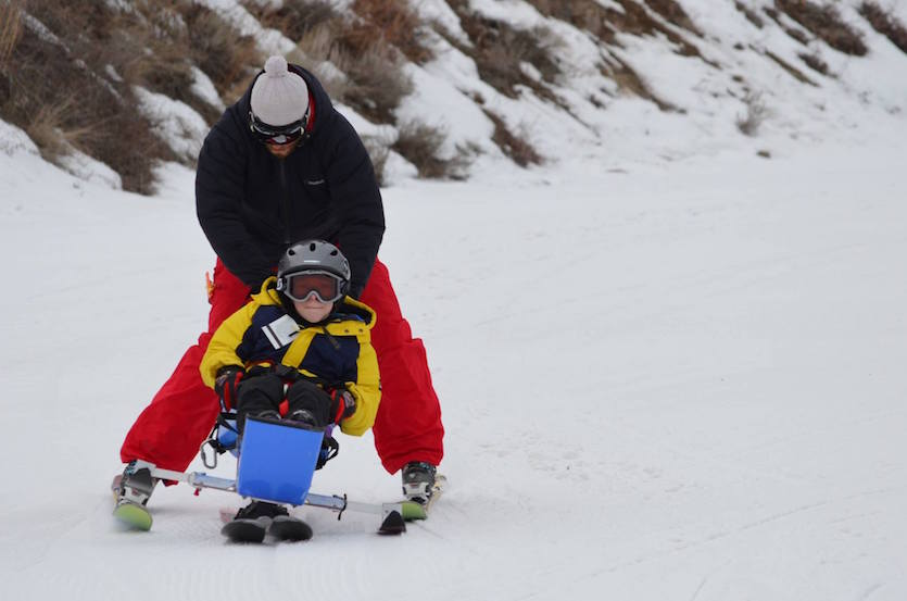 A photo of an adult skier with a child adaptive skier he is holding up from behind.
