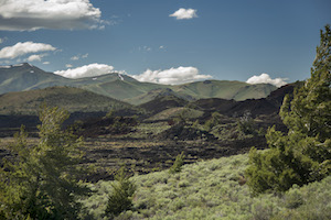 A scenic picture of the Craters of the Moon National Monument with green vegetation in the foreground, black-rock lava flows in the middle and mountains, clouds and sky in the background.