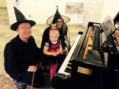 Two people dressed as witches and a young girl, all sitting at a piano.